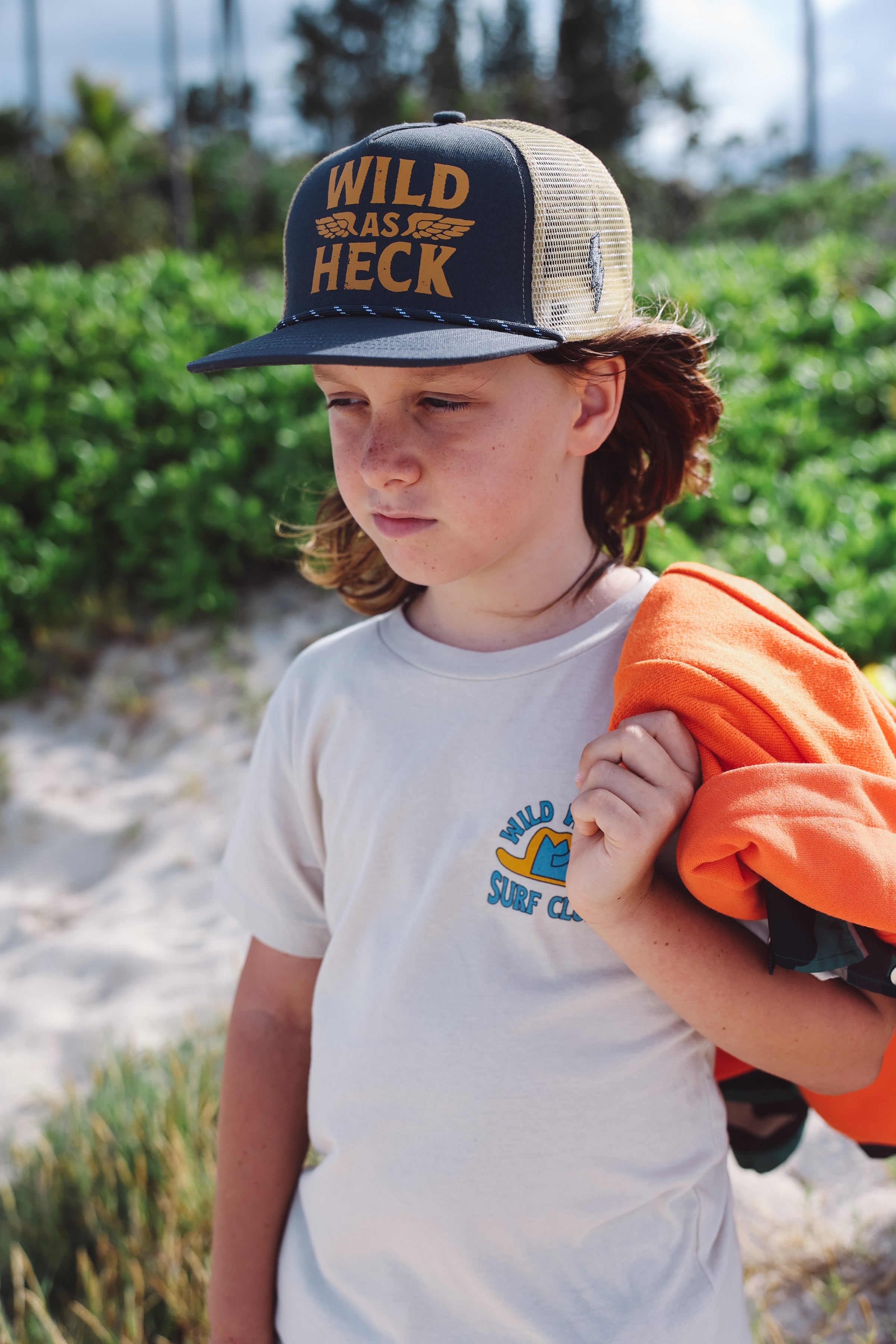 Child wearing a 'Wild as Heck' cap and holding an orange shirt on a beach.