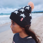 Child wearing a black star-patterned beanie with a pink pom-pom on a beach