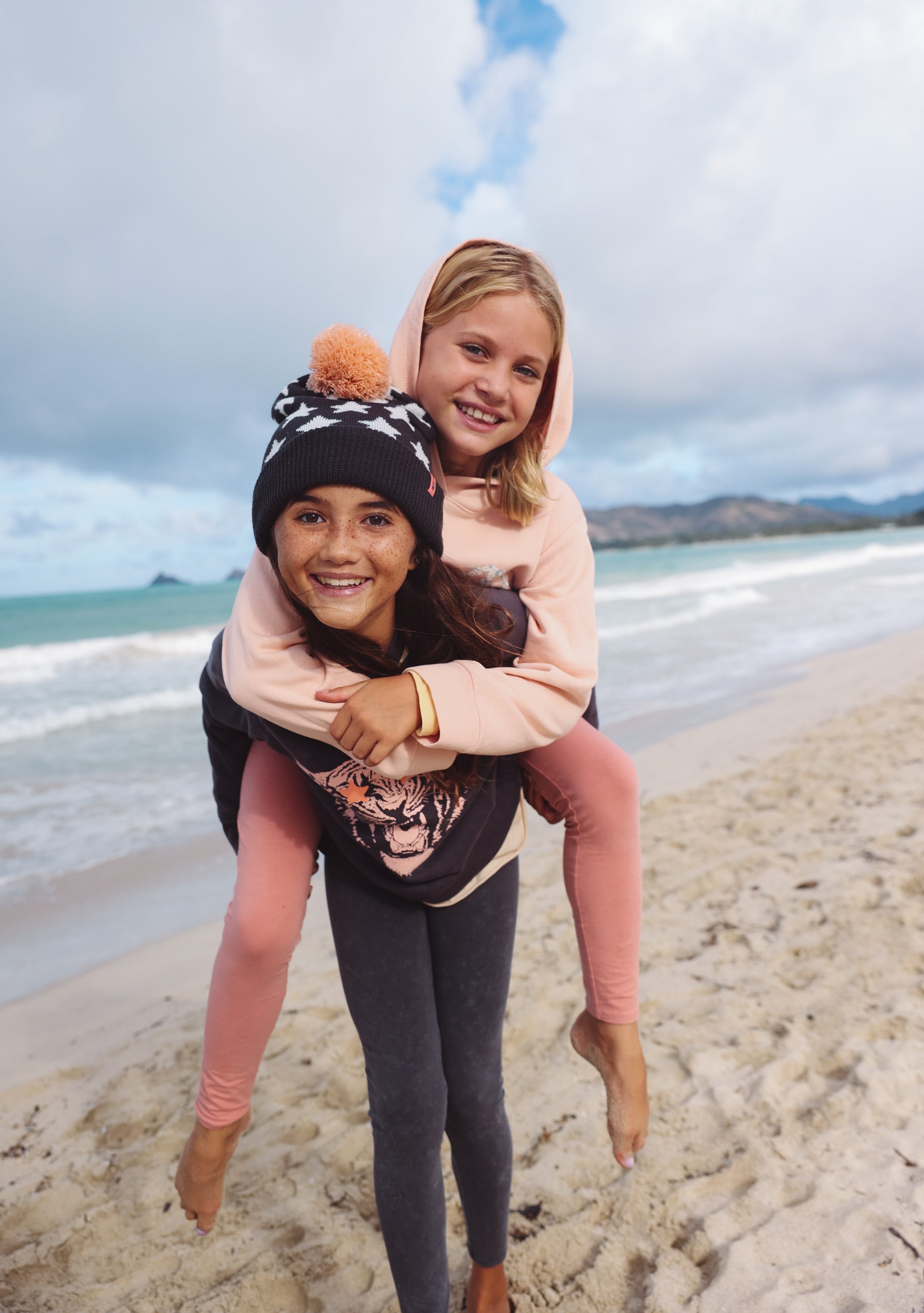 Two young girls on a beach with one girl piggybacking the other, smiling at the camera.