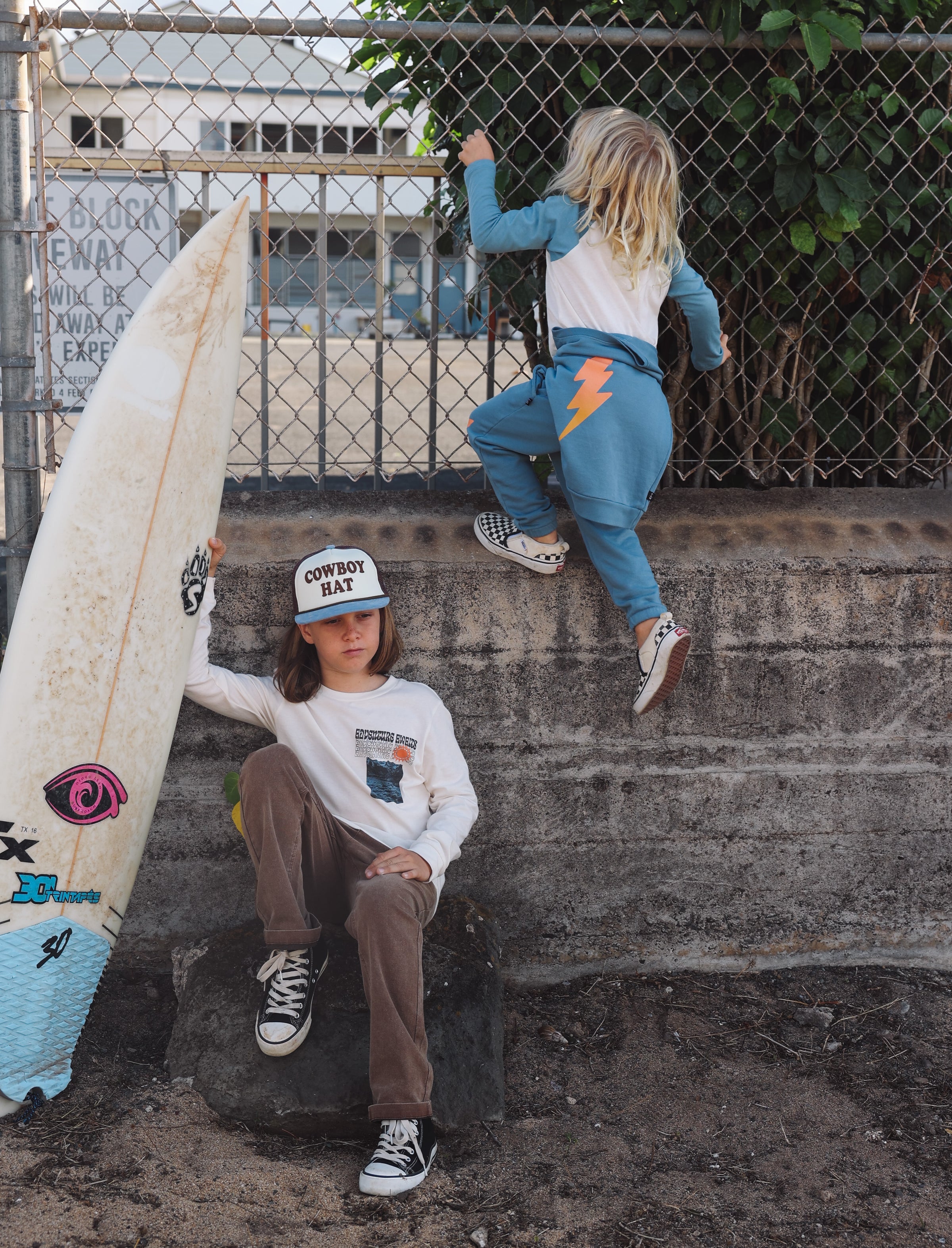 Two children with a surfboard on a concrete surface with a metal fence and building in the background.