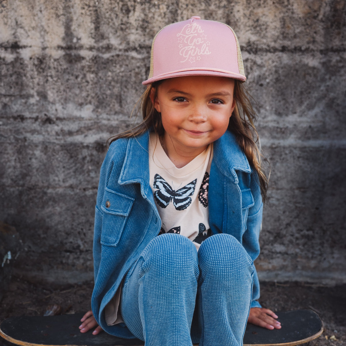 Child wearing a pink cap and blue coat sitting on a skateboard against a stone wall.