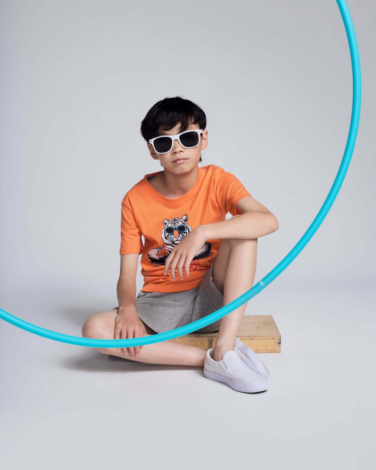 Child wearing an orange t-shirt with a graphic design, sitting on a wooden block with a blue hula hoop.