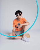 Child wearing an orange t-shirt with a graphic design, sitting on a wooden block with a blue hula hoop.