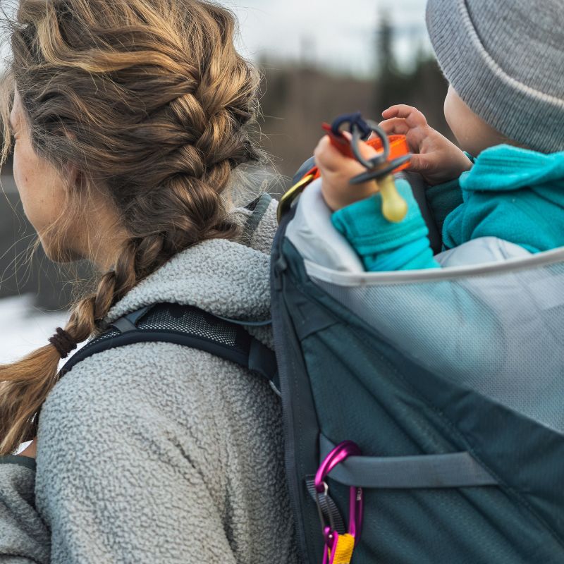 Woman with braided hair wearing a gray jacket, carrying a child in a teal jacket on her back.