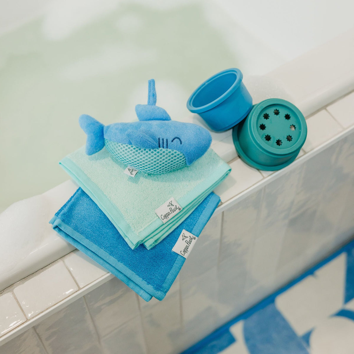 Set of blue bath toys including a shark-shaped toy and two cups on a bathtub ledge.