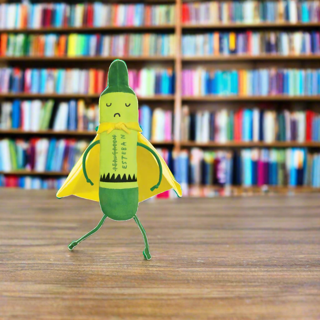 A small green and yellow toy figure resembling a stylized banana with a cape stands on a wooden table in front of blurred bookshelves.
