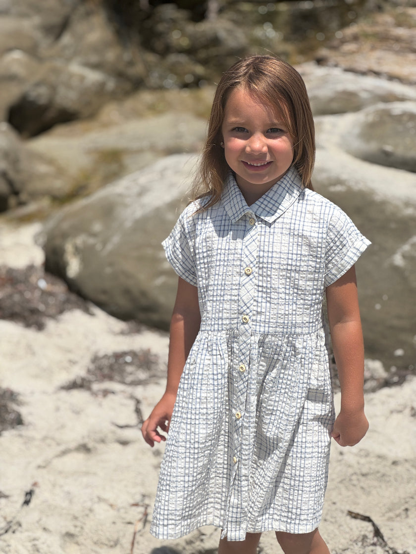 Young girl in a checkered dress standing on a rocky beach.