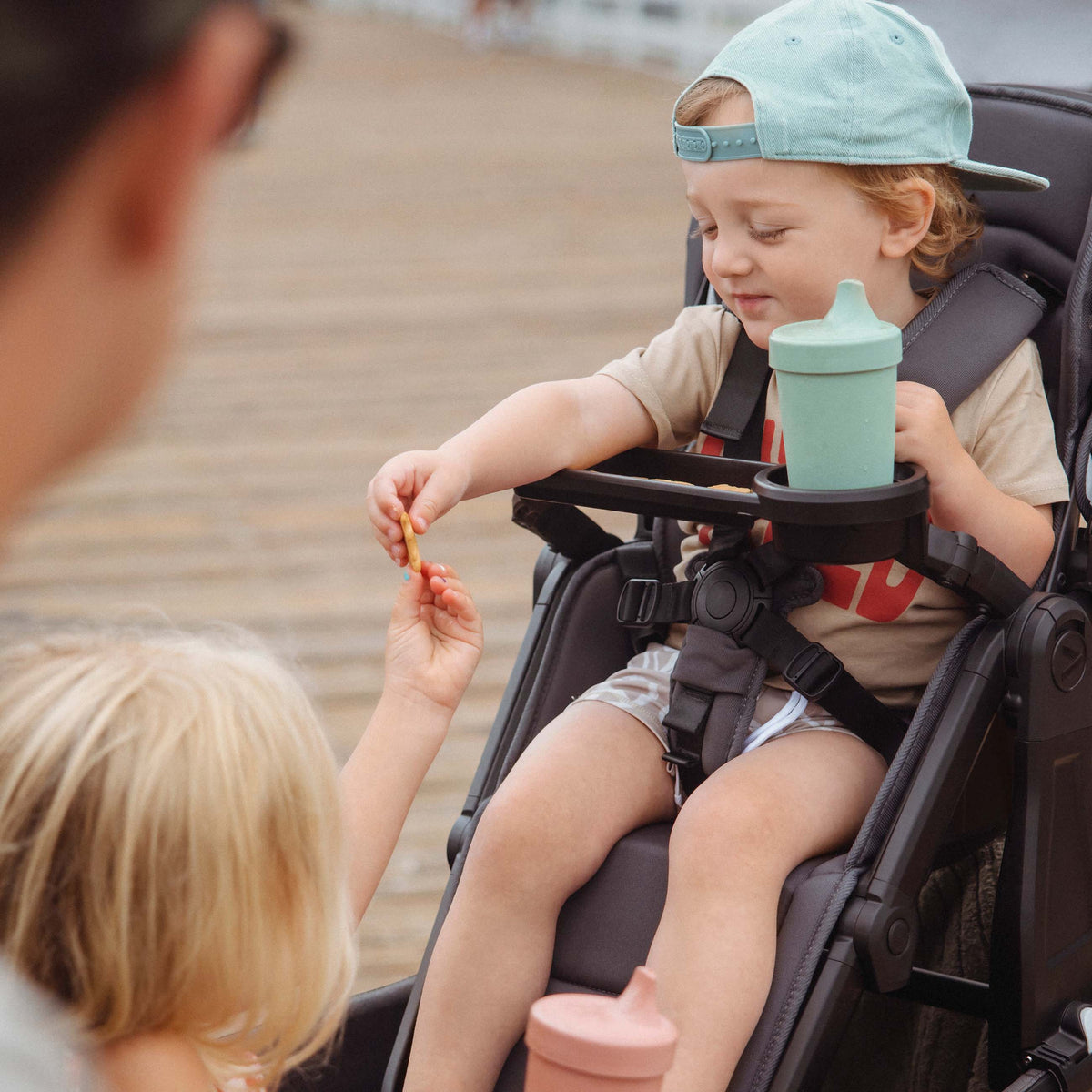 Child in a high chair with sippy cups, interacting with another child outdoors.
