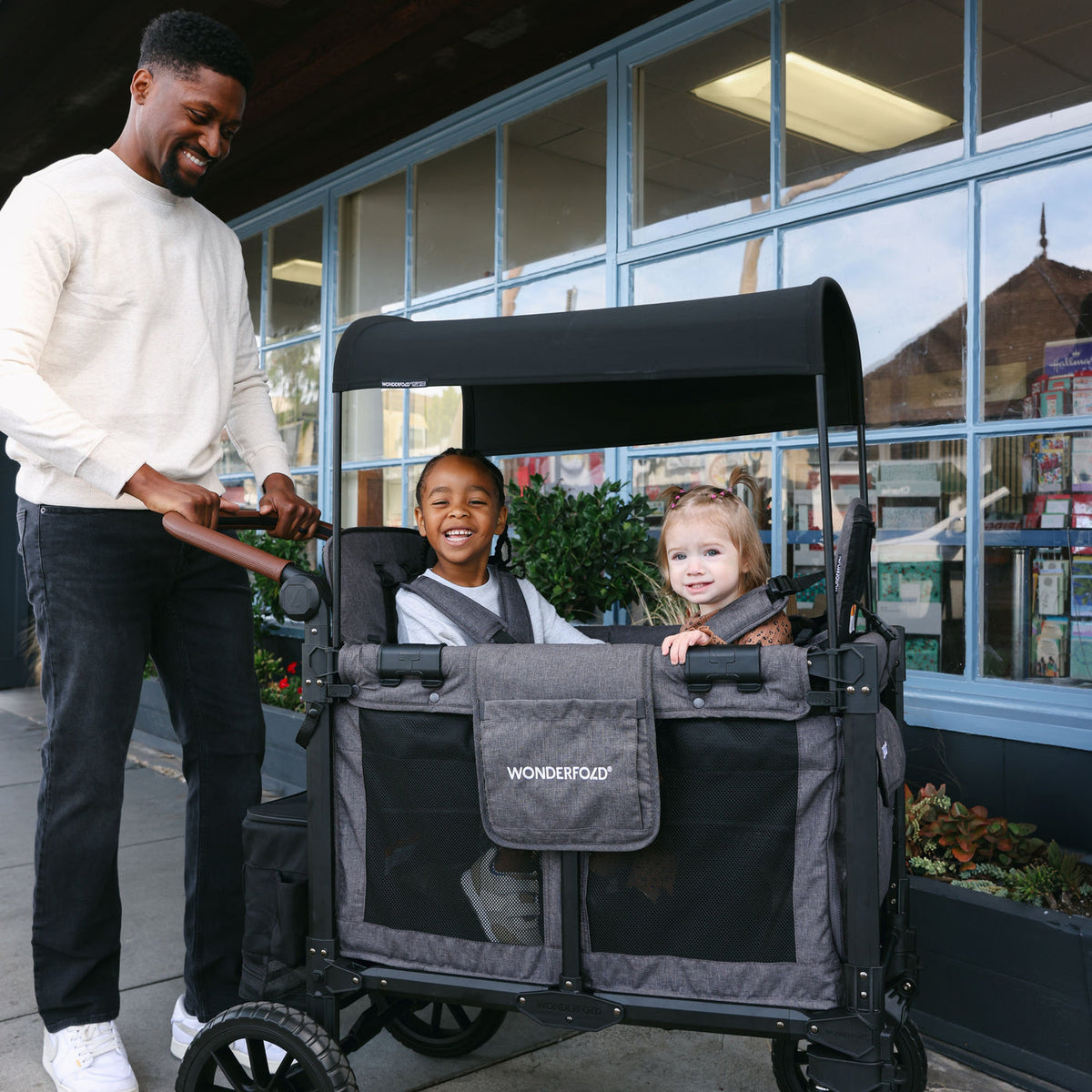Man pushing a stroller with two children in front of a store.