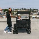 Woman pushing a black wagon with a child inside on a concrete surface.