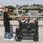 Woman pushing a black WonderFold stroller with two children on a waterfront boardwalk.