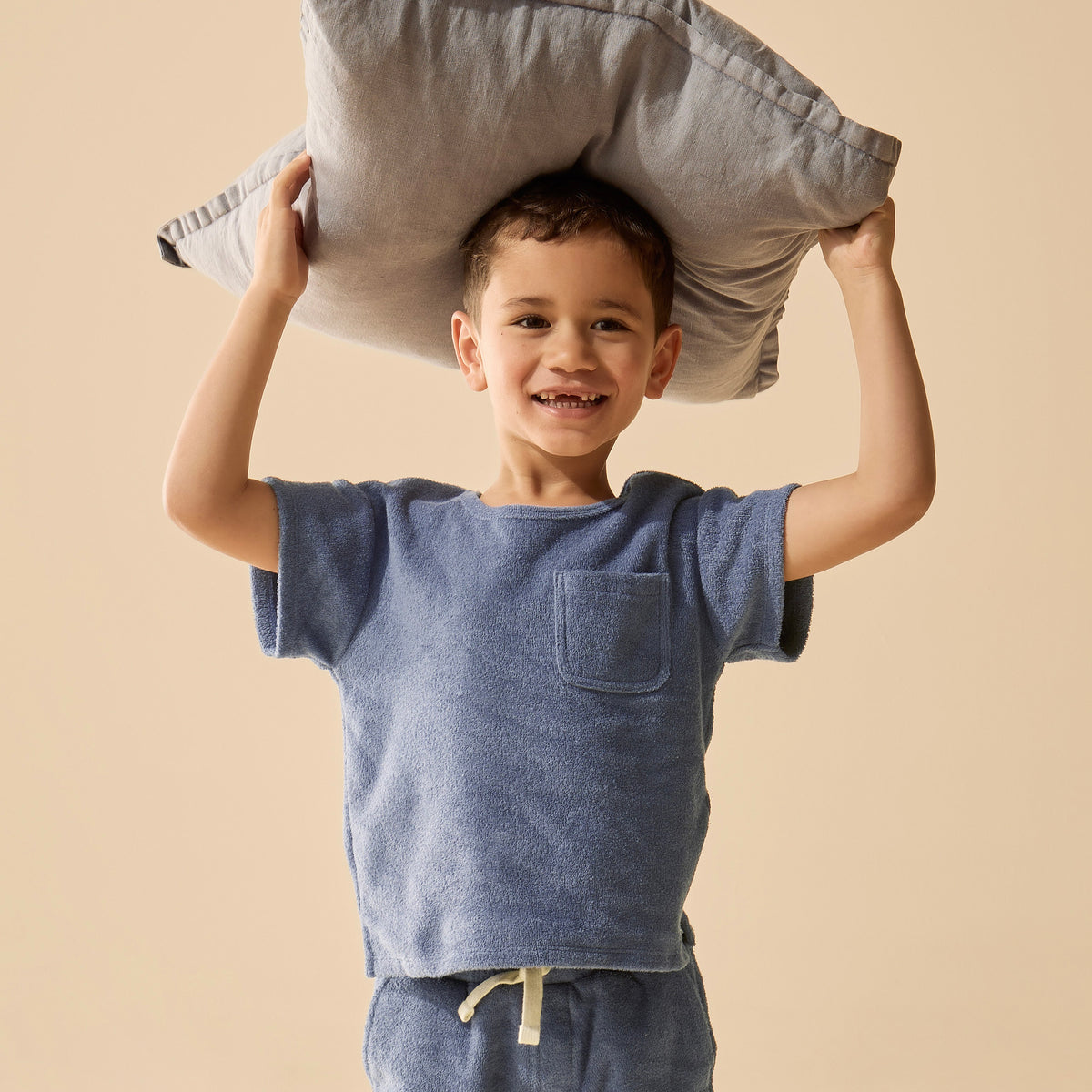 Child wearing a blue shirt and shorts, holding a gray pillow above their head against a beige background