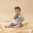Baby sitting on a beige blanket wearing a light blue shirt with text and gray shorts.