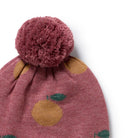 A close-up of a knit pom-pom beanie in dusty rose pink with a yellow and green fruit pattern, featuring a large textured pom-pom on top, set against a plain white background.