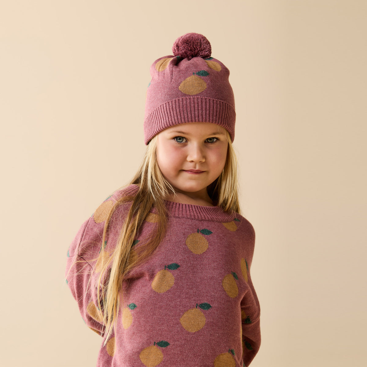 Young girl wearing a pink sweater and hat with fruit patterns on a beige background