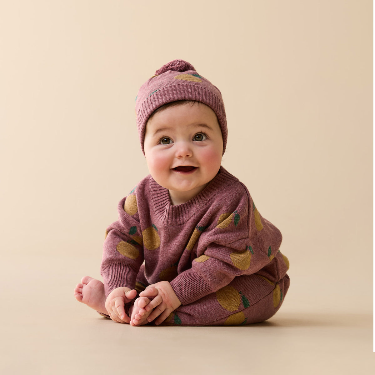 A smiling baby sitting on a beige studio background, wearing a matching knit hat and long-sleeve top in dusty mauve with scattered olive-green and mustard yellow fruit motifs.