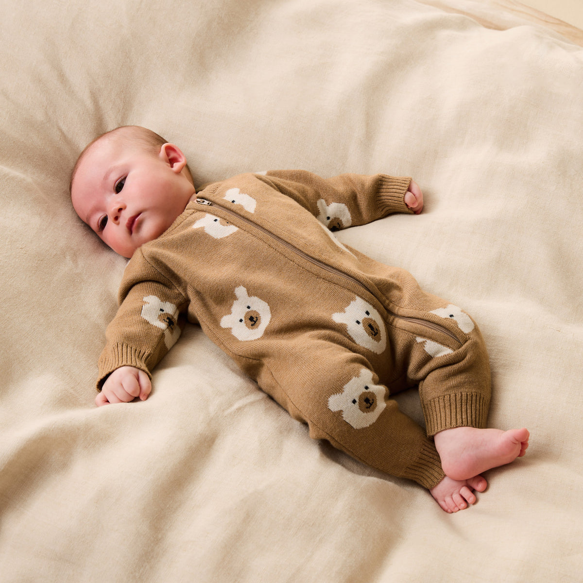 Baby wearing a brown onesie with bear patterns lying on a beige blanket