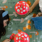 Two children playing with red paper clocks on a green tablecloth with mushroom patterns.