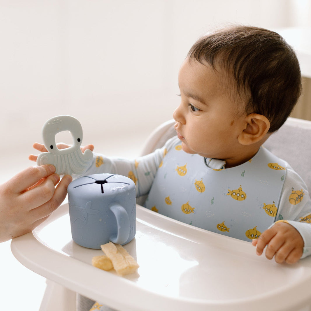 Baby in a high chair with a teething toy and bib, holding a small cup.