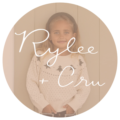 Blond 4 year old girl with a headband and a rylee & cru sweater that is cream with green fir trees standing in front of a beige wall