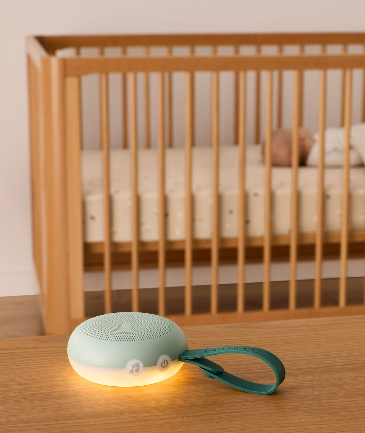 Mint green baby monitor with a glowing base on a wooden surface, with a crib and baby in the background.