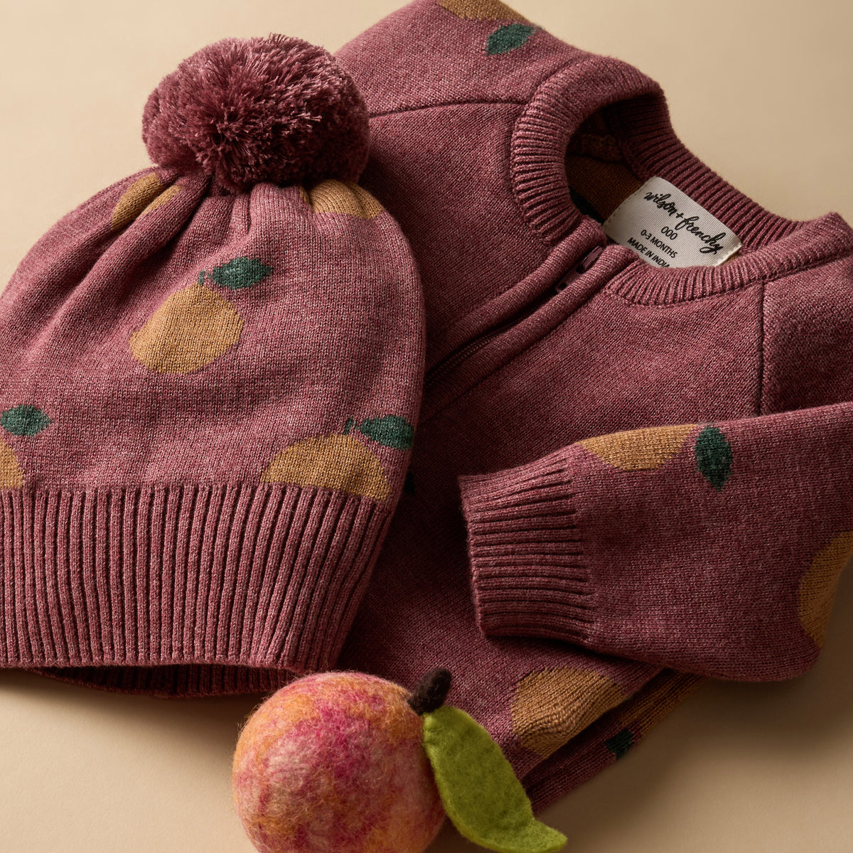 A cozy knit children's sweater and matching beanie set in dusty rose, decorated with a yellow pear pattern and green leaf accents, styled with a felt pear on a warm beige background.