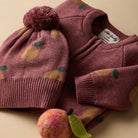 A cozy knit children's sweater and matching beanie set in dusty rose, decorated with a yellow pear pattern and green leaf accents, styled with a felt pear on a warm beige background.