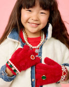 A young girl smiling while wearing plush red mittens with jeweled and pearl trim, a red sweater, a pearl necklace, and a denim jacket against a pink background.