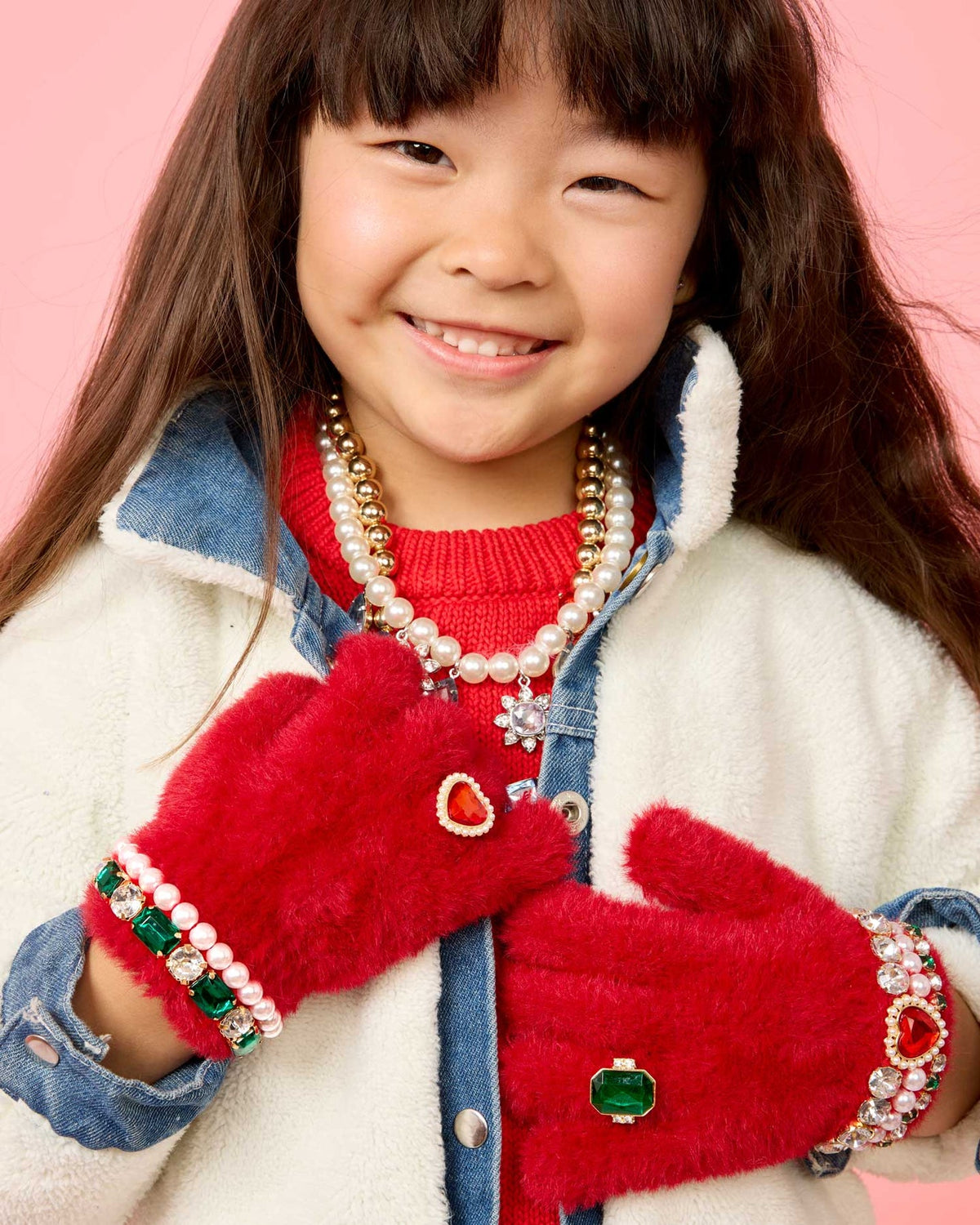 A young girl smiling while wearing plush red mittens with jeweled and pearl trim, a red sweater, a pearl necklace, and a denim jacket against a pink background.