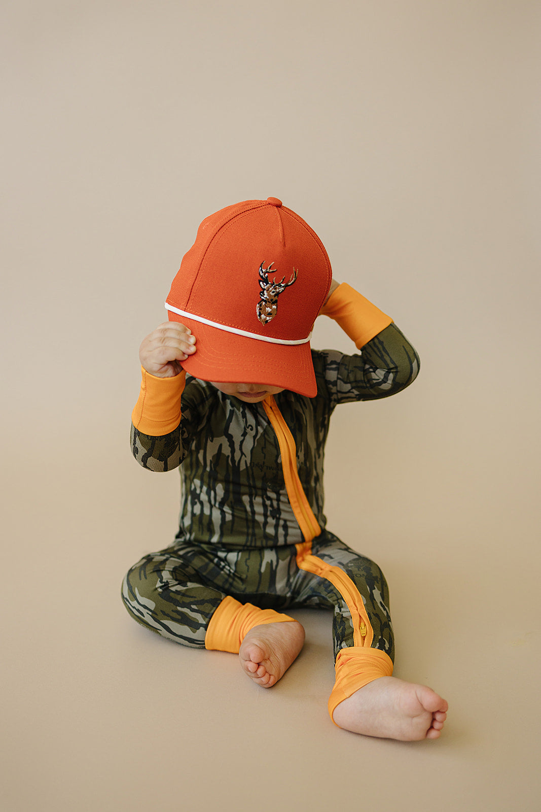 Child wearing a camouflage outfit with orange accents and an orange cap on a beige background