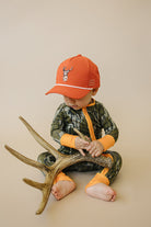 Child wearing an orange cap and camouflage outfit sitting on a large deer antler against a beige background