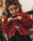 A young child wearing a red knit cardigan with silver bow details, standing in a softly blurred indoor setting.
