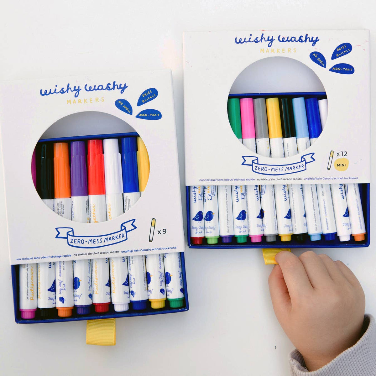 Two boxes of Wishy Washy zero-mess markers on a white background with a child's hand reaching towards one of them.