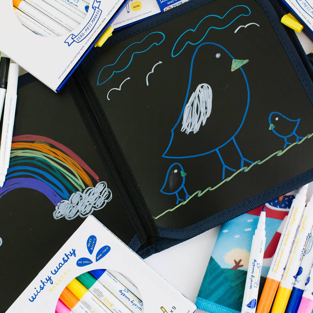 Black drawing board with colorful drawings of birds and a rainbow, surrounded by markers and coloring books.