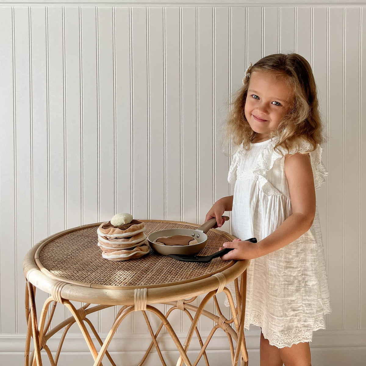 Young girl in a white dress standing next to a small round table with a stack of plates and a bowl.