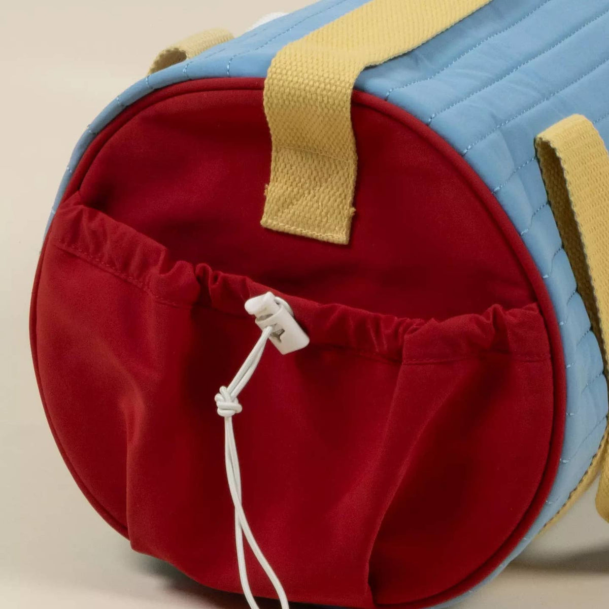 A close-up of a cylindrical duffel bag with a light blue quilted body, a red drawstring pouch on the front, and beige webbing handles.