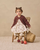 A toddler girl sits on a rustic wooden chair in a styled studio setting, wearing a cream dress with a cherry print and a dark brown cardigan. Red apples are scattered on the beige textured rug next to a small wooden basket.