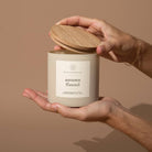 Hand holding a candle labeled 'autumn flannel' with a wooden lid against a beige background