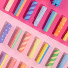 A top-down view of pastel-colored cylindrical sticks, likely crayons or wax sticks, arranged diagonally on a split pink and magenta background. The sticks feature various patterns including stripes, polka dots, swirls, and solid colors in shades of blue, yellow, green, orange, purple, and pink.