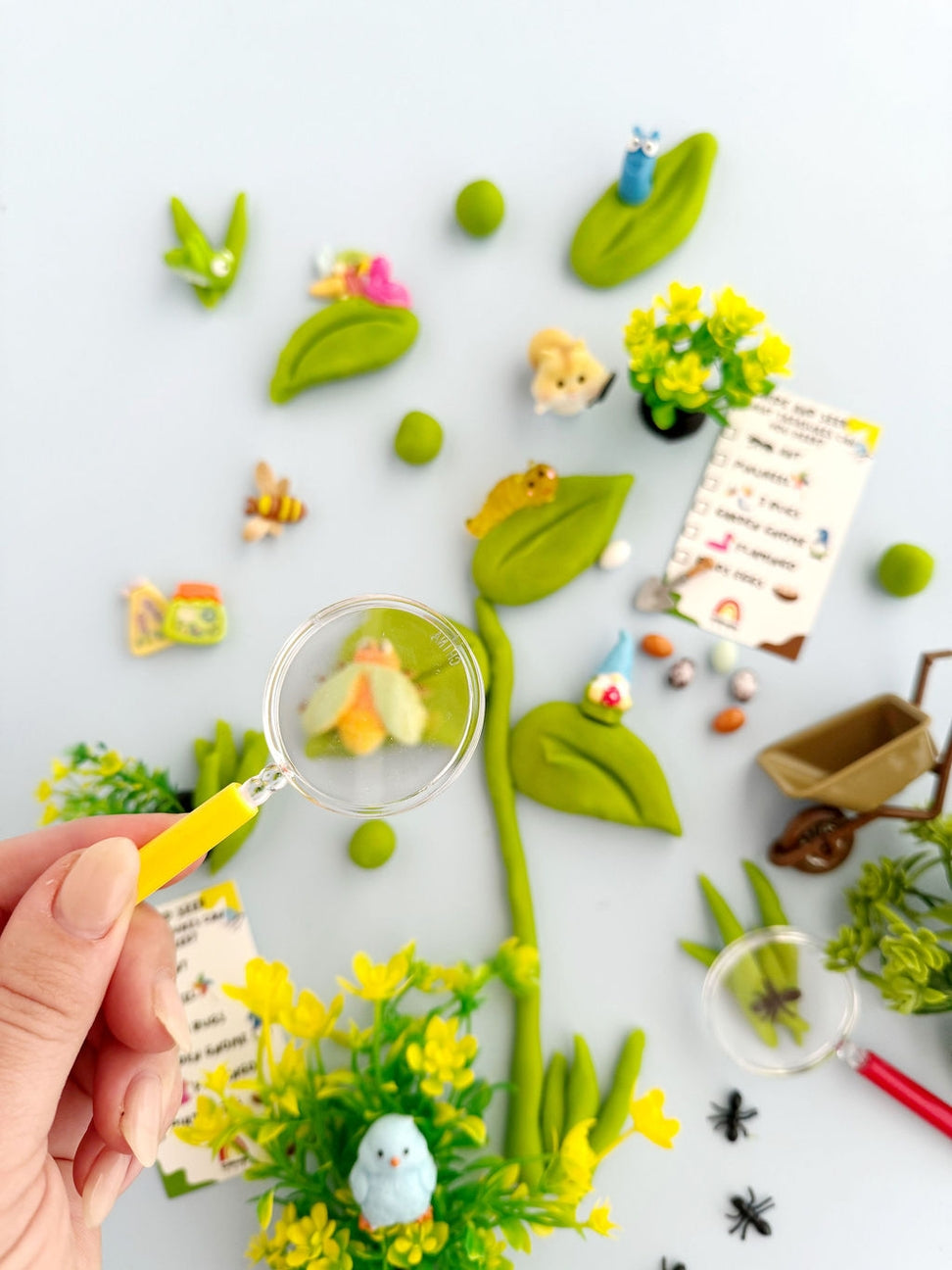 Playful garden scene with a hand holding a magnifying glass, surrounded by small plants, flowers, and animals on a white background.
