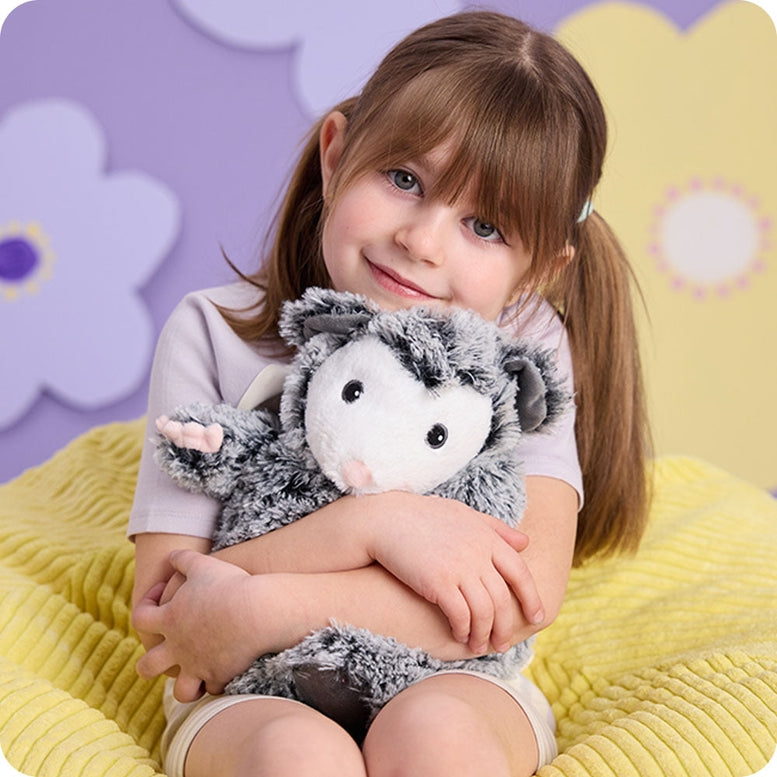A young girl with bangs and pigtails smiling while warmly hugging a gray and white fluffy Opossum by Warmies stuffed animal against a light-colored background with flowers.