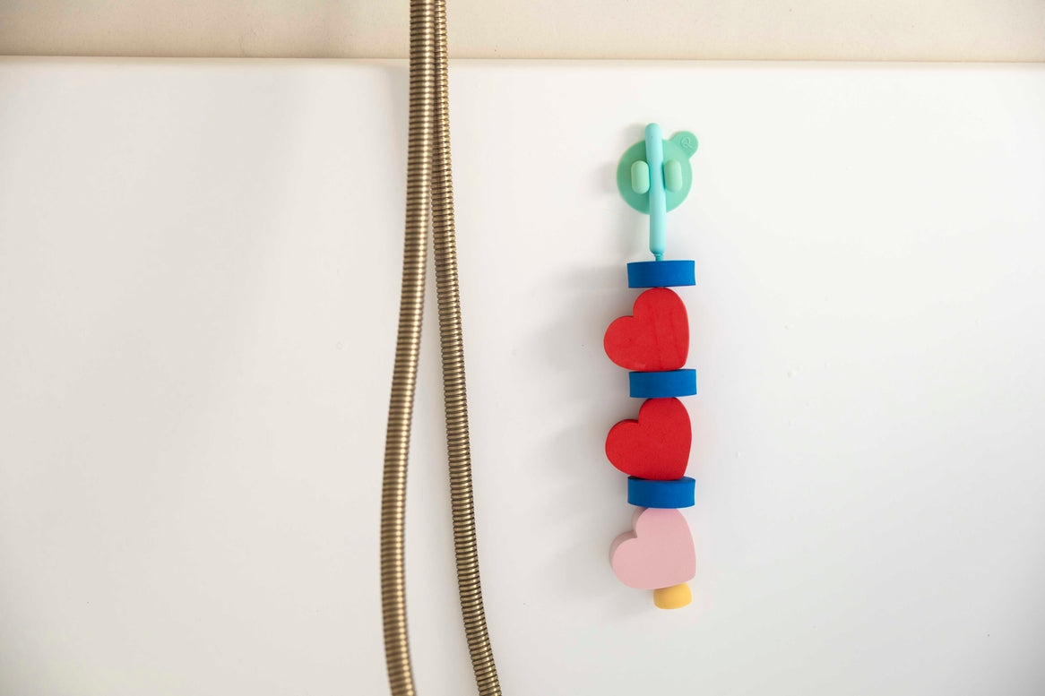 A colorful shower hook with a mint green dollar sign design and stacked red, blue, pink, and yellow heart shapes, hanging on a white bathtub next to a gold shower hose.