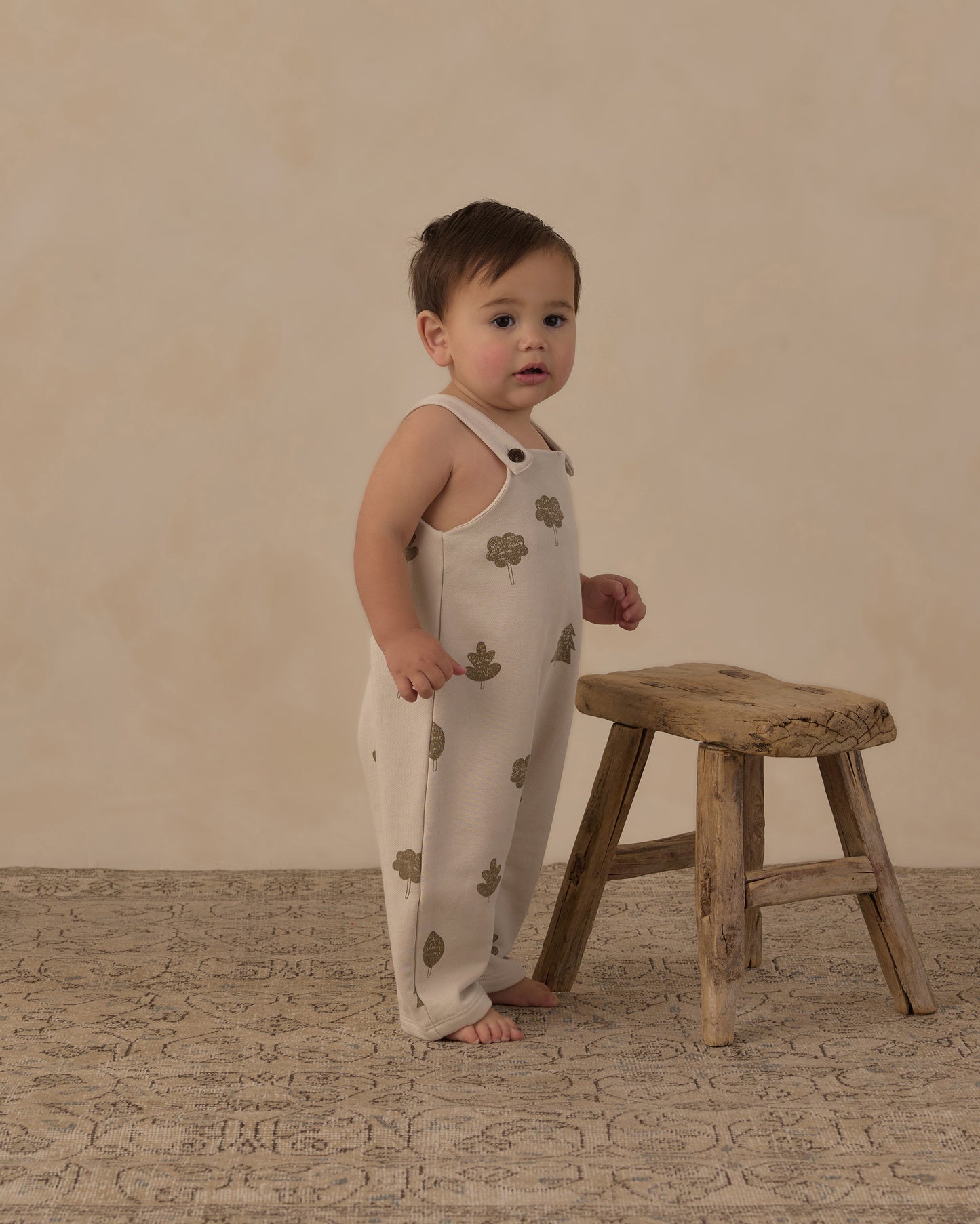 A baby standing barefoot next to a rustic wooden stool, wearing a cream-colored sleeveless jumpsuit with a brown tree-leaf pattern, set against a warm beige textured backdrop.
