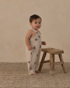 A baby standing barefoot next to a rustic wooden stool, wearing a cream-colored sleeveless jumpsuit with a brown tree-leaf pattern, set against a warm beige textured backdrop.