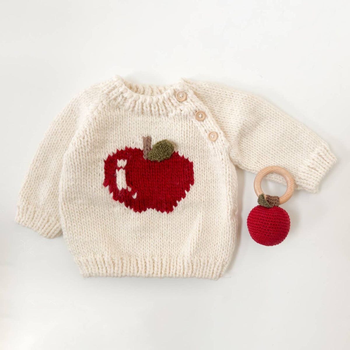 Knitted baby sweater with apple design and wooden teething ring on a light background