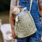 Person holding a green quilted bag with a doll inside, wearing blue overalls.