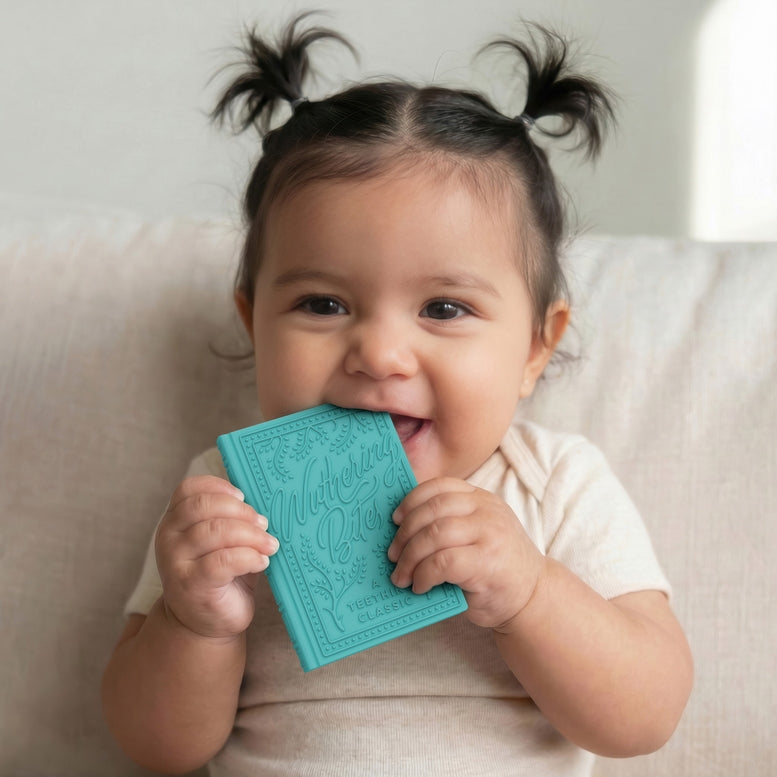 A baby with dark hair in two pigtails smiles while holding a turquoise embossed book-shaped silicone pacifier clip to their mouth, sitting on a light beige fabric couch.
