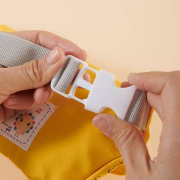 Close-up of hands adjusting a white plastic buckle on a yellow fabric with a cartoon character label.