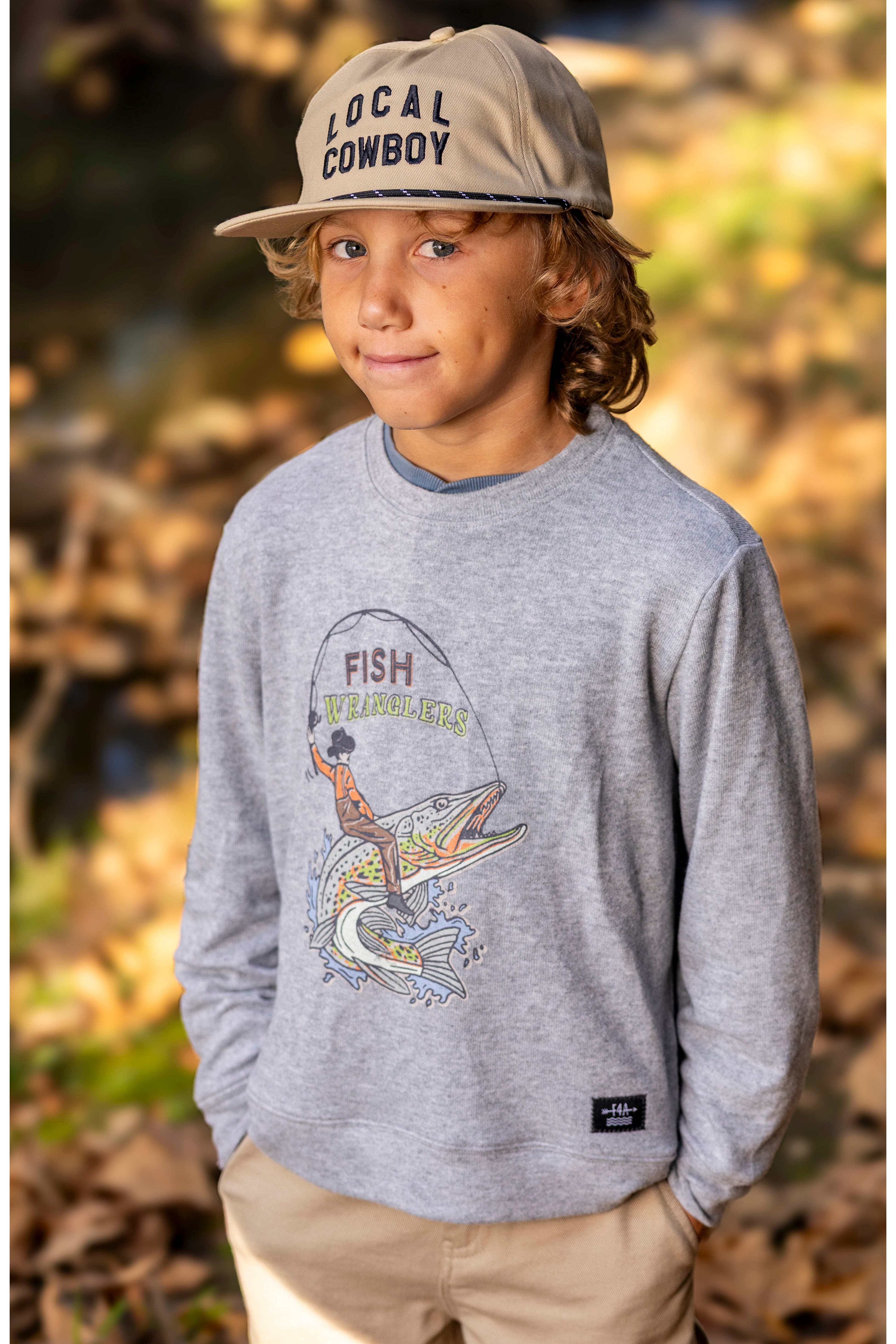 A young boy wearing a heather gray long-sleeve graphic sweatshirt with a fisherman graphic on the front, khaki pants, and a beige baseball cap that says 'LOCAL COWBOY'. He is standing outdoors in a wooded setting with fallen leaves.