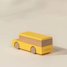 A wooden toy bus by Cocoon Block, featuring a bright yellow painted body and natural wood wheels and side panel, set against a warm beige background.